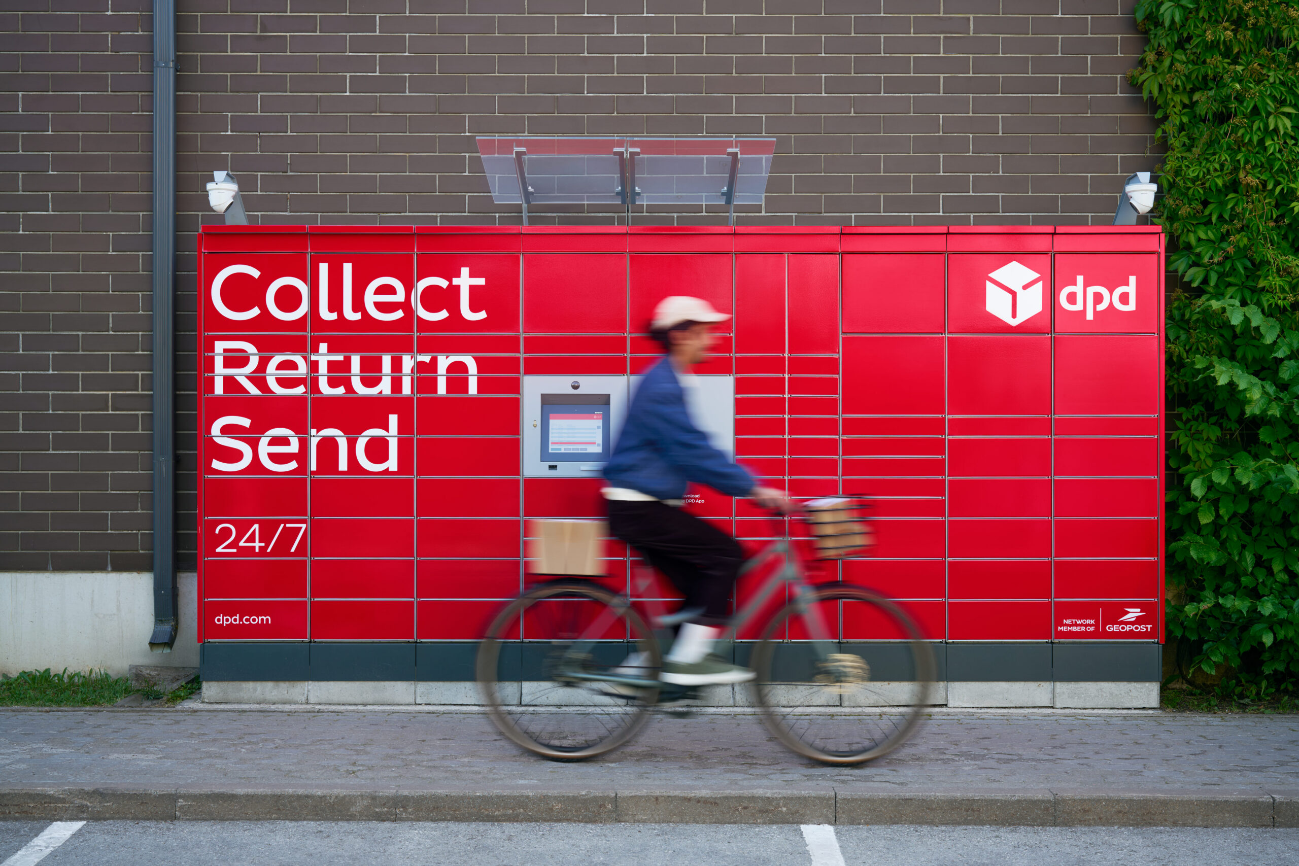 Red parcel locker in the background, branded with a dpd logo, woman on a bike passing by